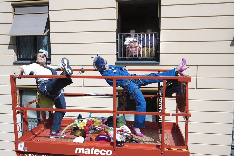 Zwei Clowns verdrehen auf der Hebebühne vor einem Fenster einer Senioreneinrichtung ihre Körper.
