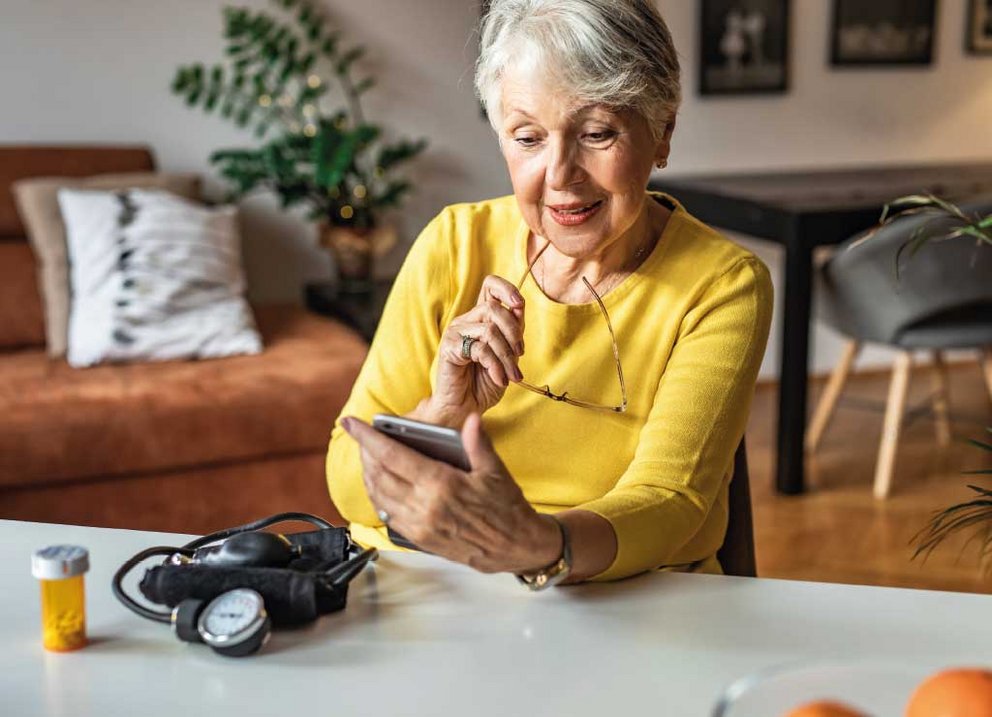 ältere Frau sieht auf Handy, neben ihr Blutdruckgerät und Tabletten