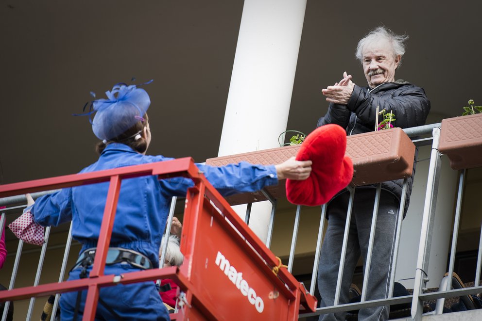 Ein Clown streckt auf einer roten Hebebühne vor dem geöffneten Fenster einer Senioreneinrichtung seine Hand nach einem älteren Herrn aus. 