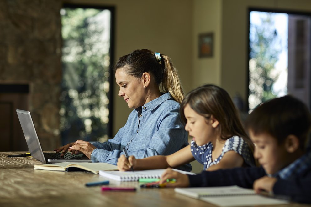 Das Bild zeigt eine Mutter mit zwei Kindern bei der Arbeit zuhause am Laptop.