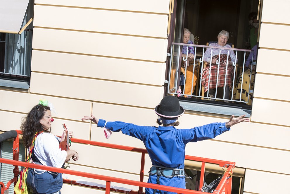 Zwei Clowns kommunizieren von einer Hebebühne aus mit zwei Senioren am geöffneten Fenster in einem Pflegeheim.