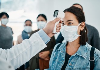 Ein Arzt hält einer Frau mit Maske im Wartezimmer ein Fieberthermometer an die Stirn.